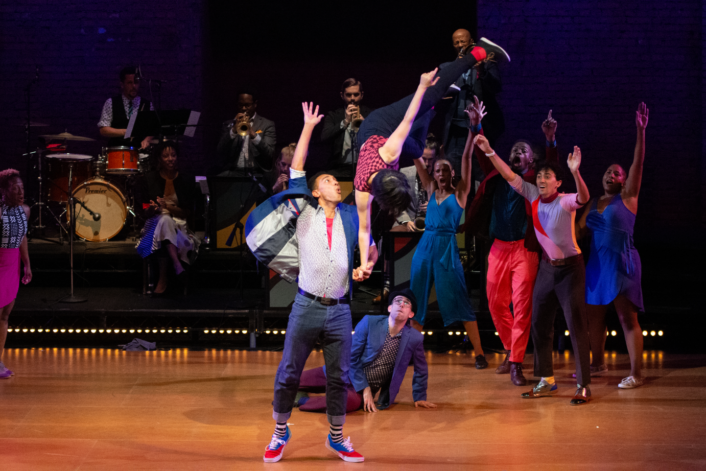 A man lifts a woman in the air with one hand. She is upside down, like in the middle of a flip. Other dancers cheer the couple on.