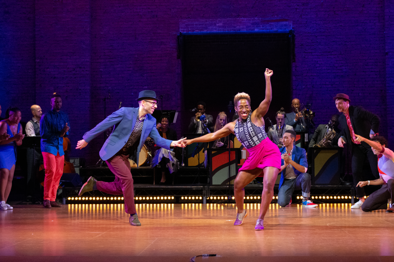 A man and a woman hold one hand as they lean into each other. They are smiling. Around them, dancers kneel in a circle.