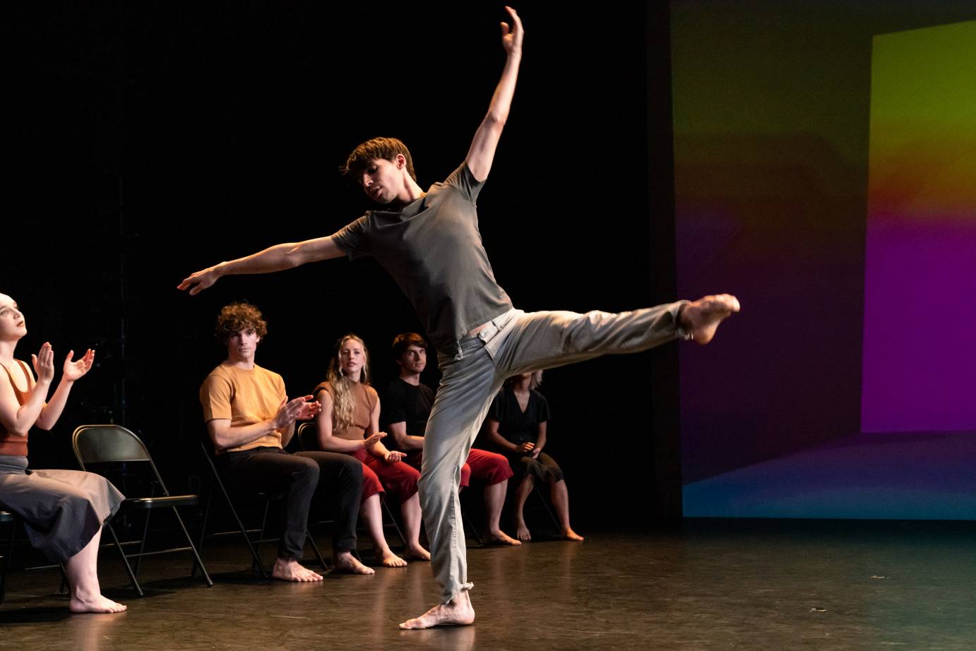 One dancer extends his leg to the side, his arms raised in a division sign. Other dancers sit on chairs in a straight line, clapping. 