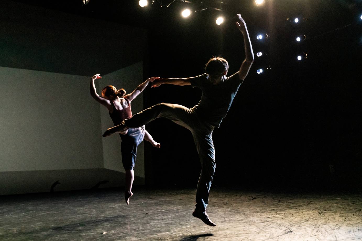 Two dancers back each other in dim light. Each has a leg lifted in a side attitude while one arms is curved overhead and the other is to the side.
